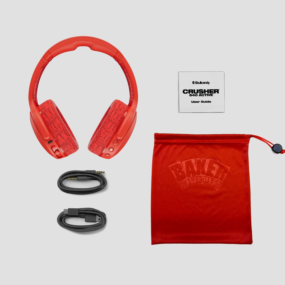 Red over-ear Crusher 540 headphones with embossed branding, shown with a black audio cable and a red drawstring pouch on a light gray background. A small card reading 'Baker Crusher Gold Active User Guide' is also visible. | Skullcandy X Baker Skateboards