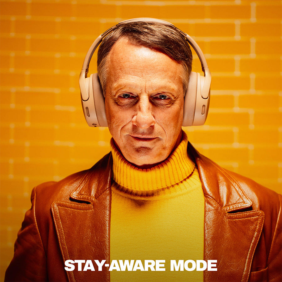 Tony hawk wearing Crusher 1080 headphones with a text 'Adjustable Stay-Aware Mode' on an orange brick wall background | Primer