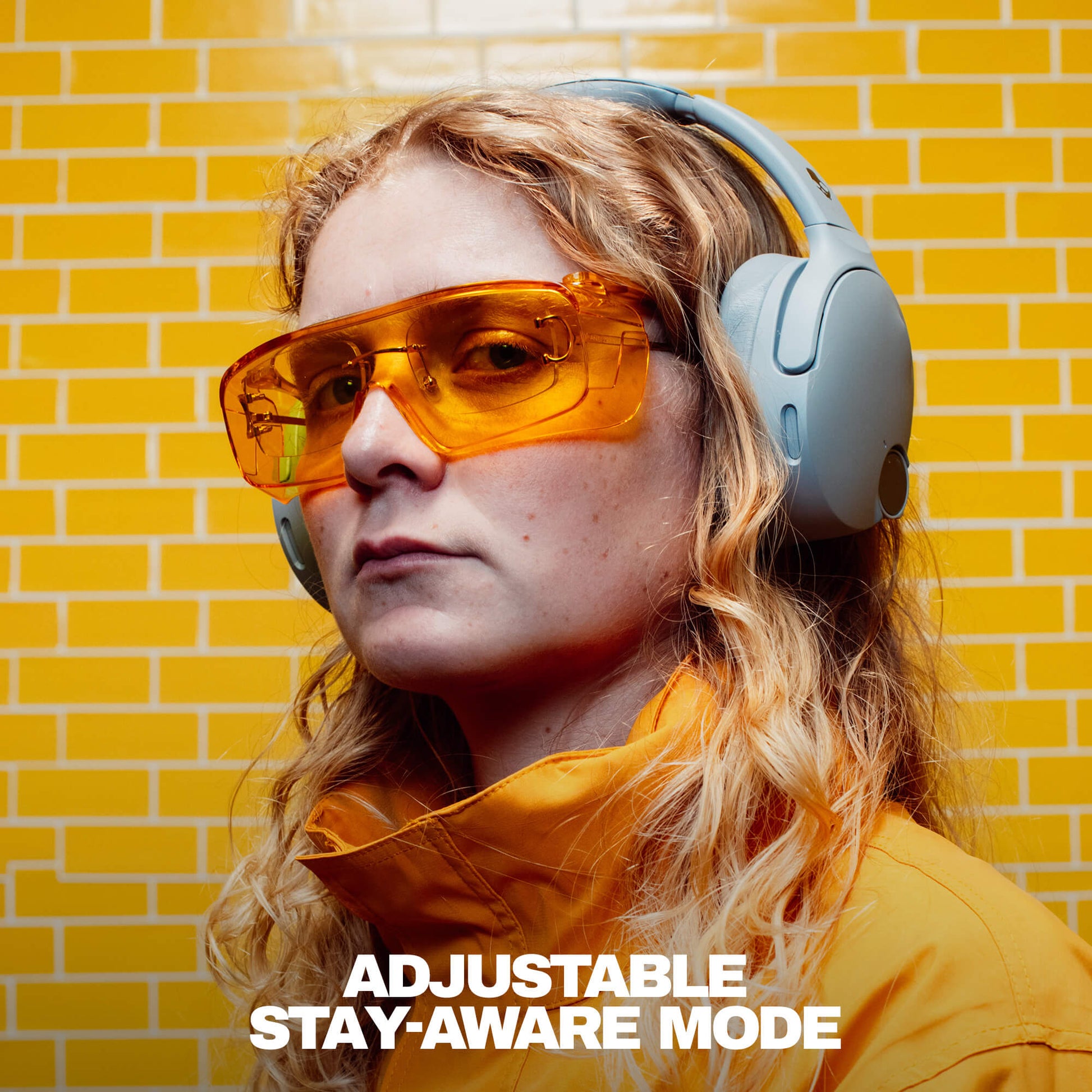 Person wearing orange sunglasses and a yellow jacket with a yellow brick wall background | Cement