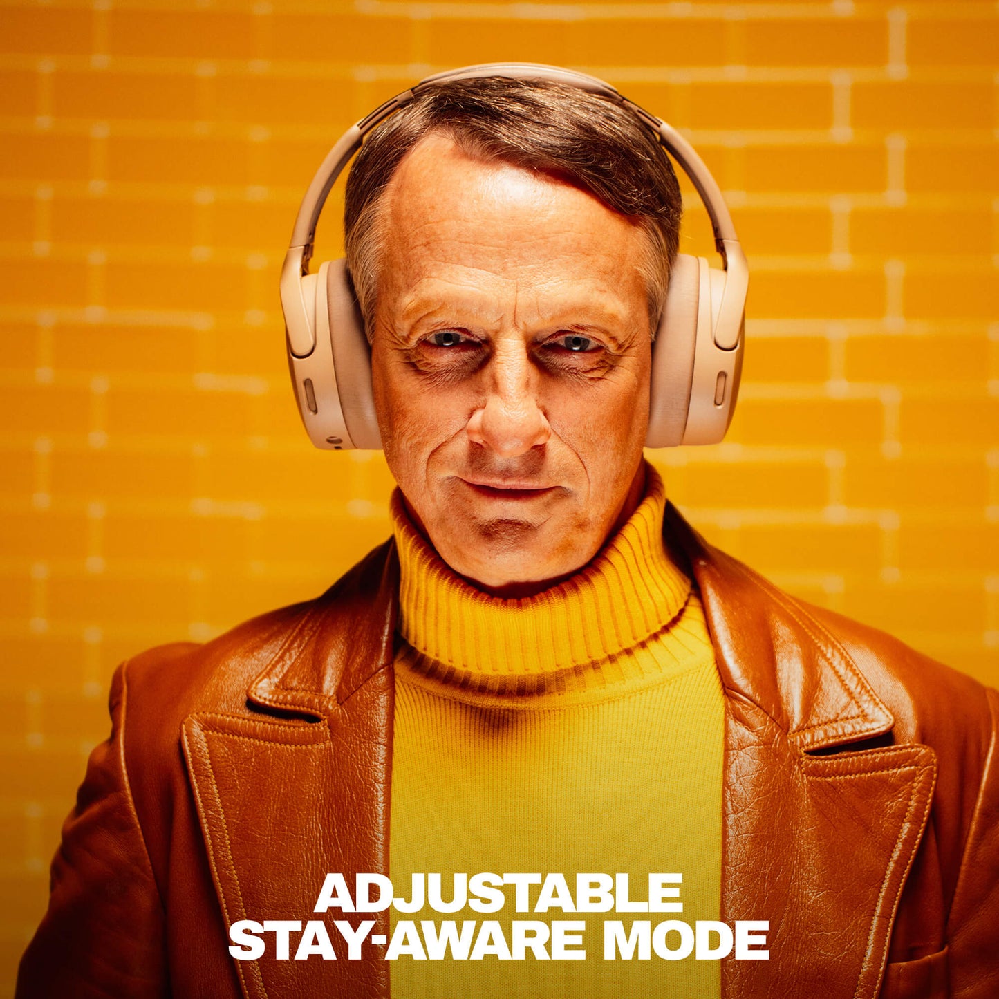 Tony hawk wearing Crusher 1080 headphones with a text 'Adjustable Stay-Aware Mode' on an orange brick wall background | Primer