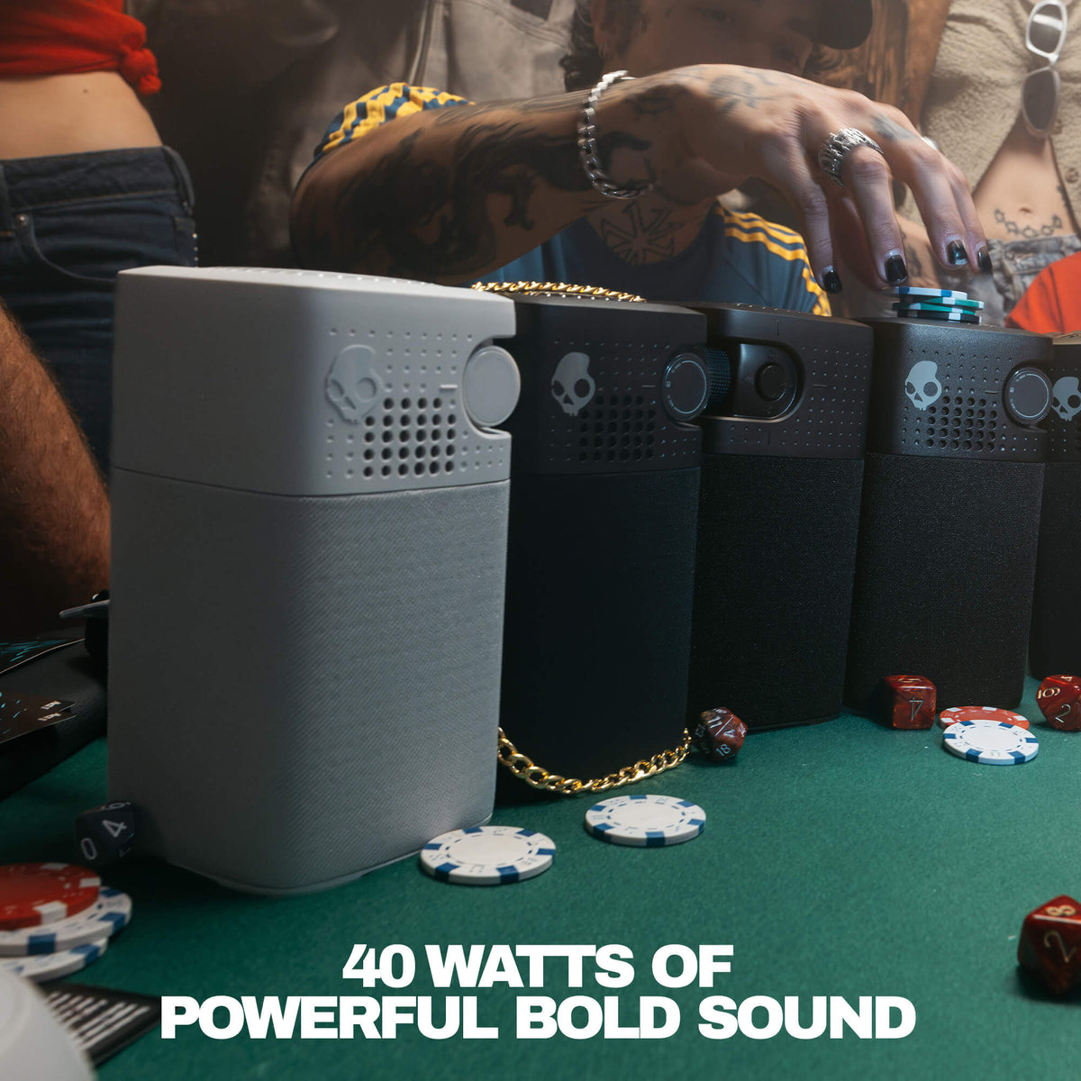 Three Skullcandy portable speakers in gray and black are lined up on a green felt table with poker chips and dice, with a person's tattooed hand reaching toward the devices. Text at the bottom reads '40 WATTS OF POWERFUL BOLD SOUND'. | True Black