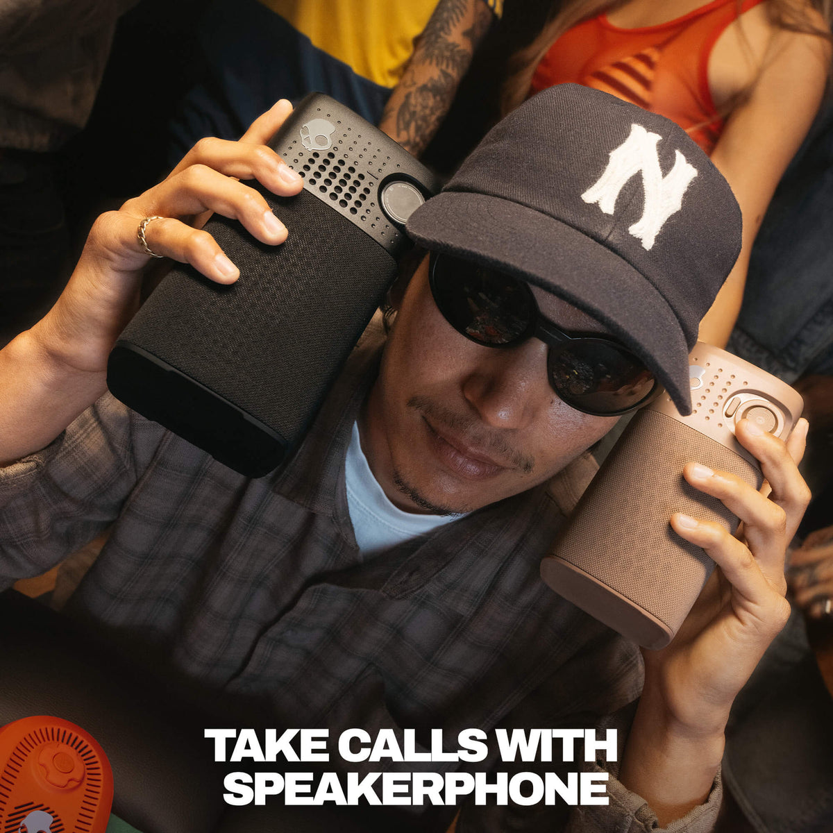 A man wearing a black baseball cap with a white 'N' logo, sunglasses, and a plaid shirt holds two session 900 speakerphones, one black and one taupe, to his ears. The text 'TAKE CALLS WITH SPEAKERPHONE' is displayed at the bottom. | Cement