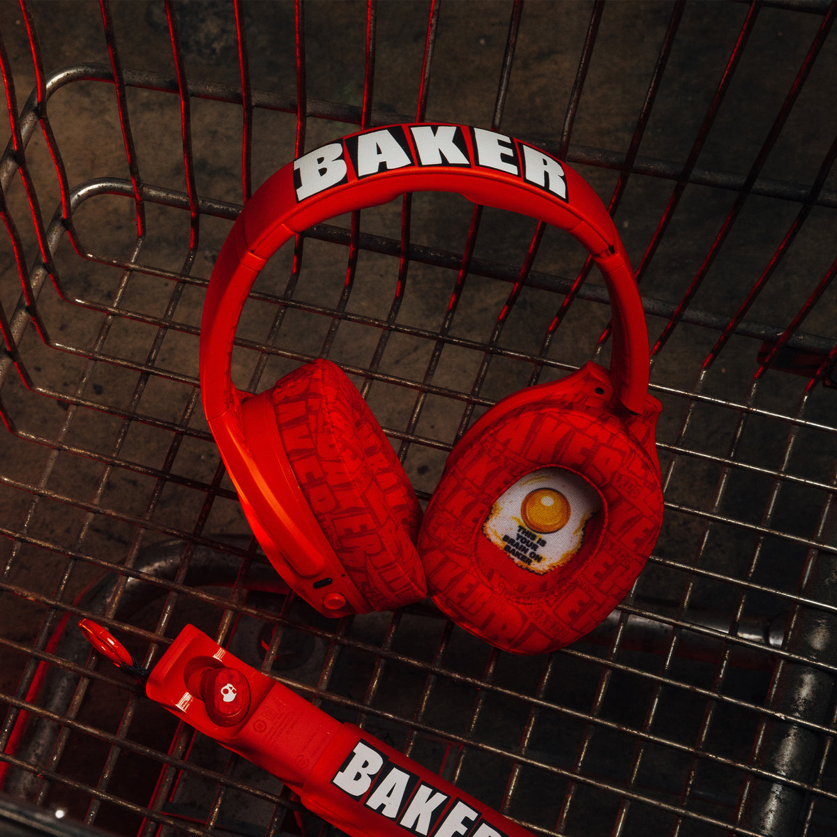 Red over-ear Crusher 540 headphones headphones with 'BAKER' printed on the headband and a matching red controller, styled inside a metal shopping cart. | Skullcandy X Baker Skateboards
