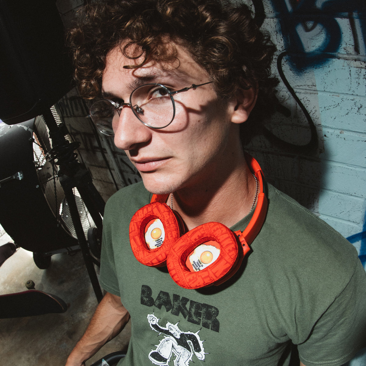 A man with curly brown hair and glasses wearing a green graphic t-shirt and red over-ear Crsuher 540 Active headphones with egg-shaped ear cushions, standing in a dimly lit studio or workshop environment. | Skullcandy X Baker Skateboards