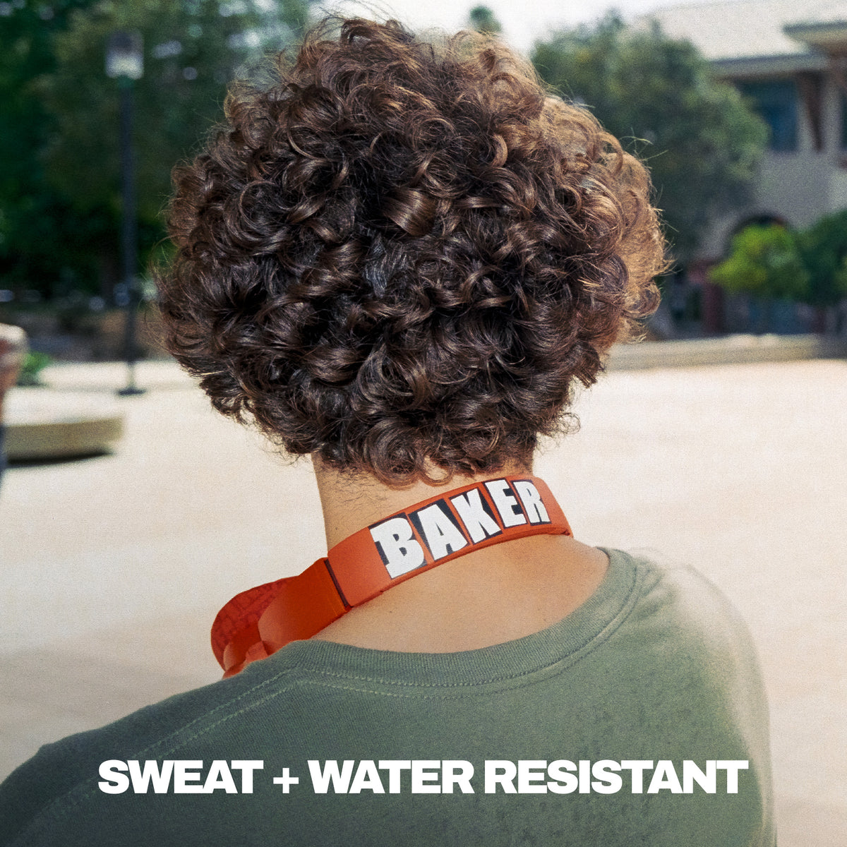 A person shown from behind outdoors wearing a bright red Crusher 540 active headphones with the name 'BAKER' printed in white. The person has curly brown hair and is wearing an olive green shirt. | Skullcandy X Baker Skateboards