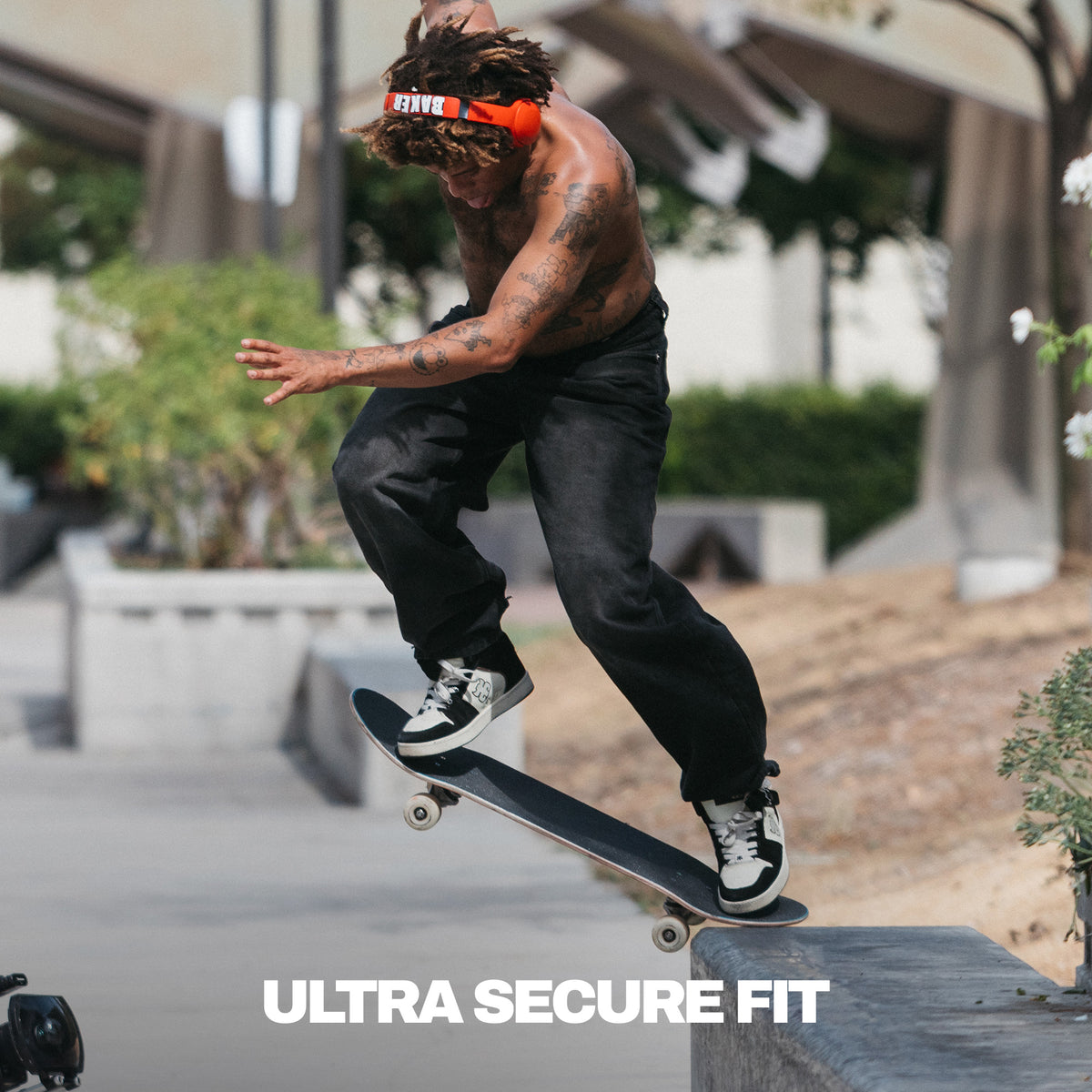 A shirtless man with tattoos performs a skateboard trick while wearing black pants, black and white sneakers, and a red pair of Skullcandy x baker skateboards crusher 540 active headphones. The text 'ULTRA SECURE FIT' is overlaid at the bottom on a concrete urban setting. | Skullcandy X Baker Skateboards