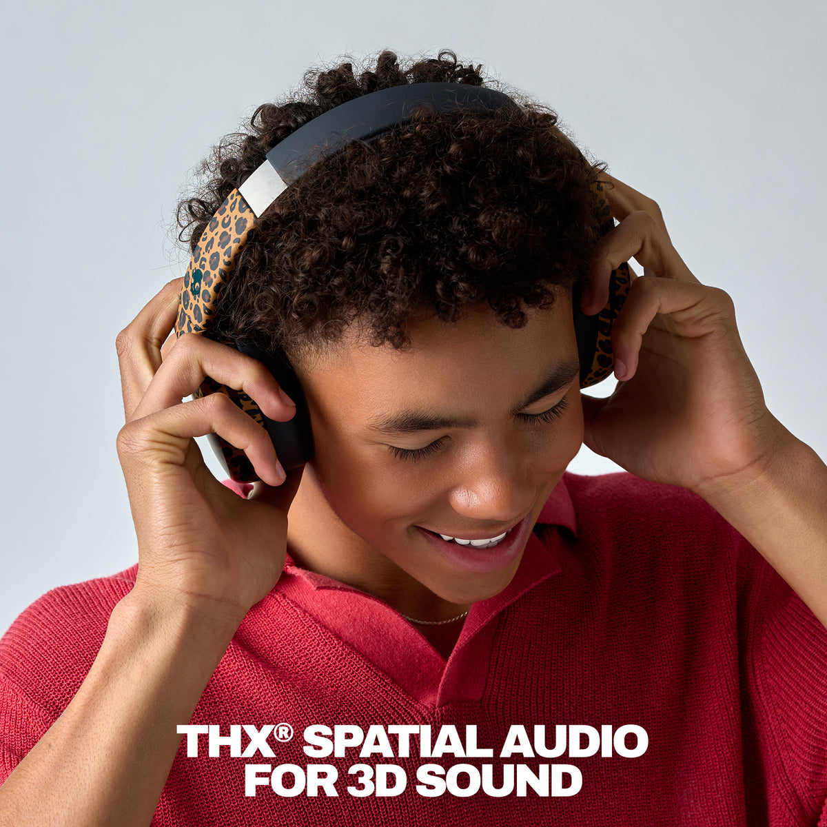 A young man with curly dark hair wearing over-ear headphones with a leopard-print pattern and a black headband, smiling while looking down. He is wearing a red knit polo shirt against a light gray studio background, with the text 'THX SPATIAL AUDIO FOR 3D SOUND' at the bottom. | Leopard
