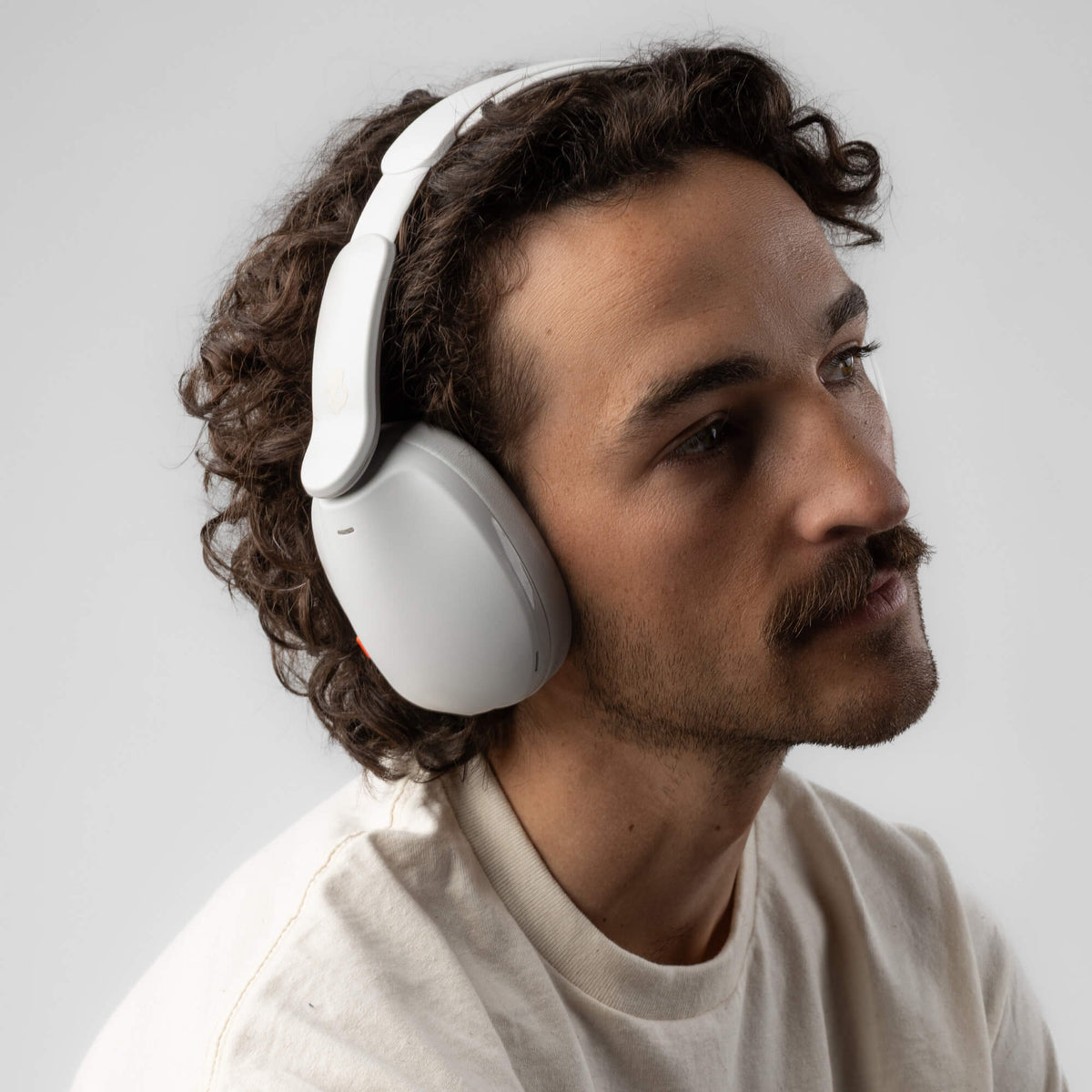 A man with curly brown hair and a mustache wearing white hesh 360 over-ear headphones, shown in a close-up profile against a light gray background. | Bone