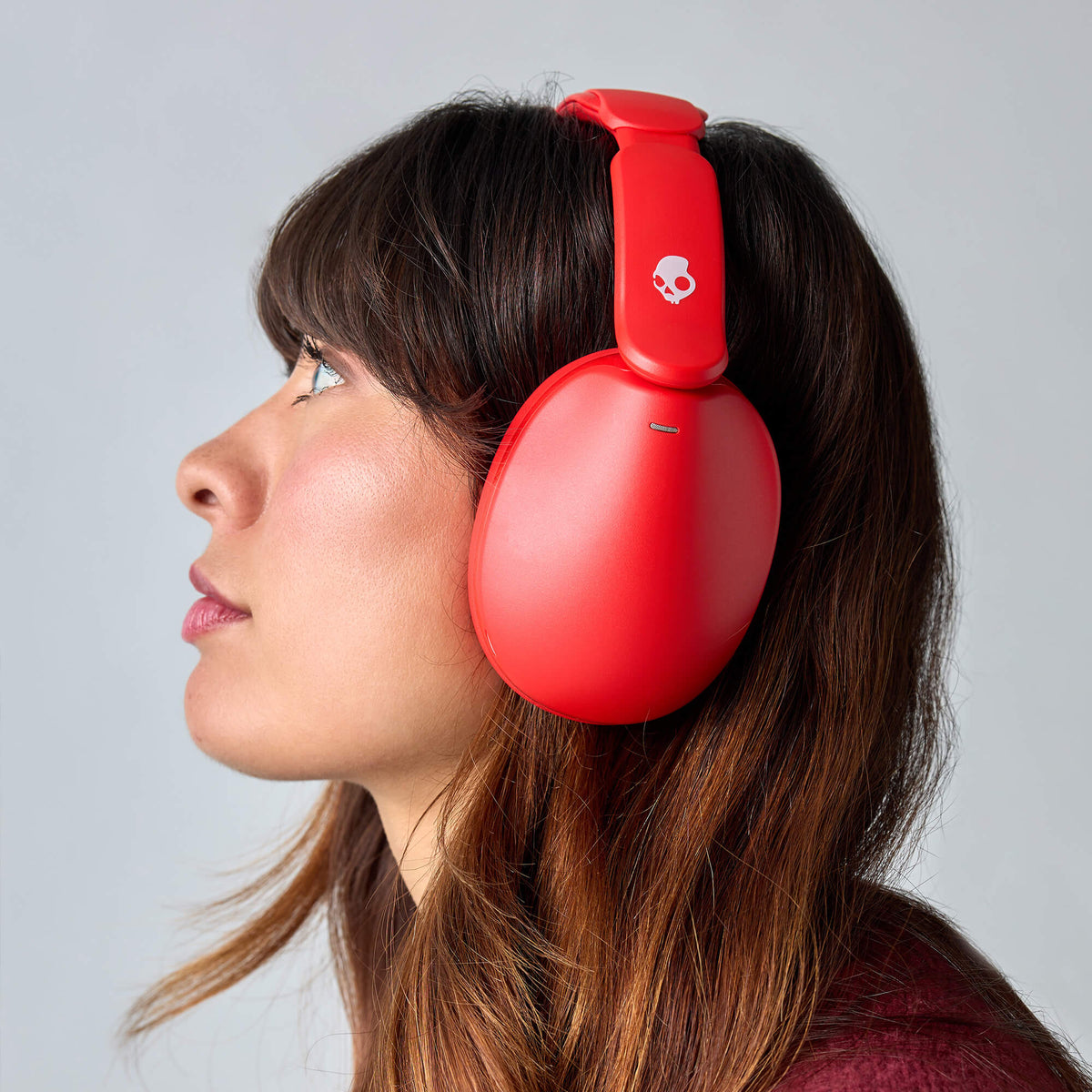 A woman in profile wearing bright red hesh 360 over-ear headphones with a small white skull logo on the headband, photographed against a light gray studio background. | Plasma