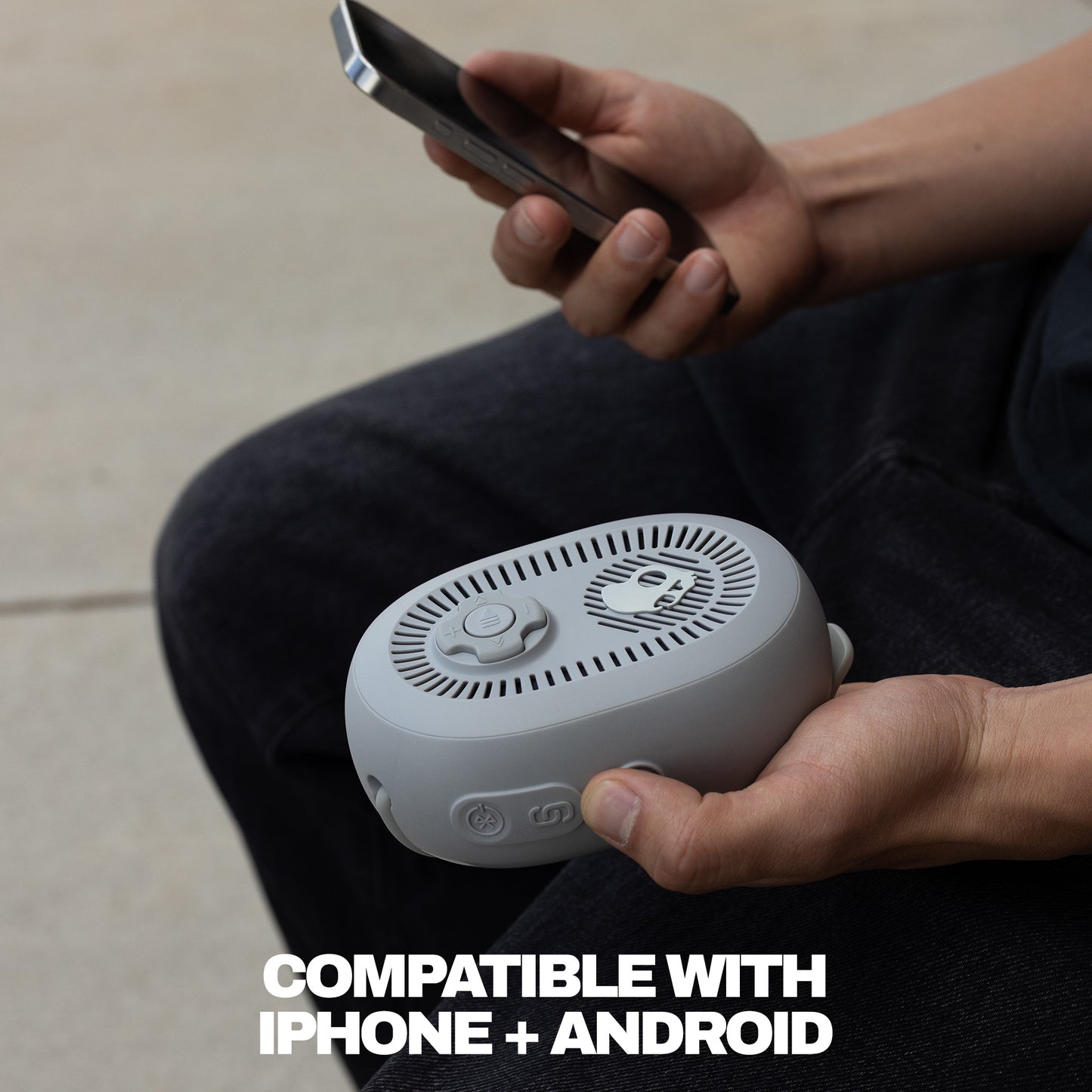Person holding a small white speaker with a phone, sitting on a light-colored floor. | Cement