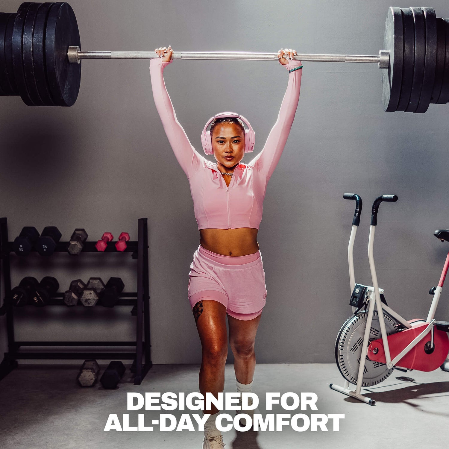 A woman in a pink athletic zip-up hoodie and matching shorts lifts a barbell overhead in a gym setting. She is wearing pink headphones, with dumbbells on a rack to her left and a stationary bike to her right.  | Candy