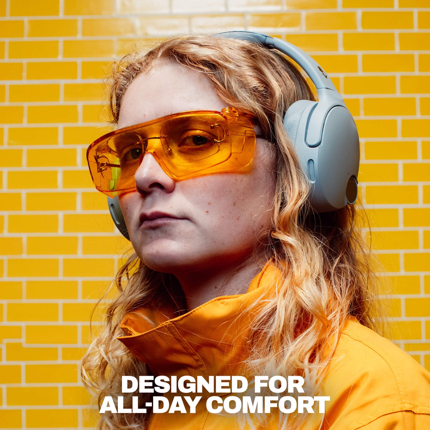 A woman wearing crusher 1080  over-ear headphones and translucent orange-tinted safety glasses, set against a yellow brick wall background with the text 'DESIGNED FOR ALL-DAY COMFORT' at the bottom. | Cement